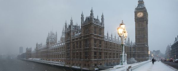 A wide winter scene showing a large historic riverside building covered in snow. The structure is ornate, with many towers, pointed spires, and rows of tall windows running along the length of the river. Snow is falling heavily, creating a hazy atmosphere that softens the outlines of the architecture. To the right, a tall clock tower rises above the rest of the scene, its clock face illuminated. In the foreground, a snow‑covered bridge spans the river, with ornate streetlamps glowing warmly. 